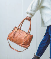 Wearing a white sweater and blue jeans, the person holds the Bed|Stü Rockababy—a compact bag crafted from vegetable-tanned leather with top handles and a crossbody strap—while standing against a light-colored wall.