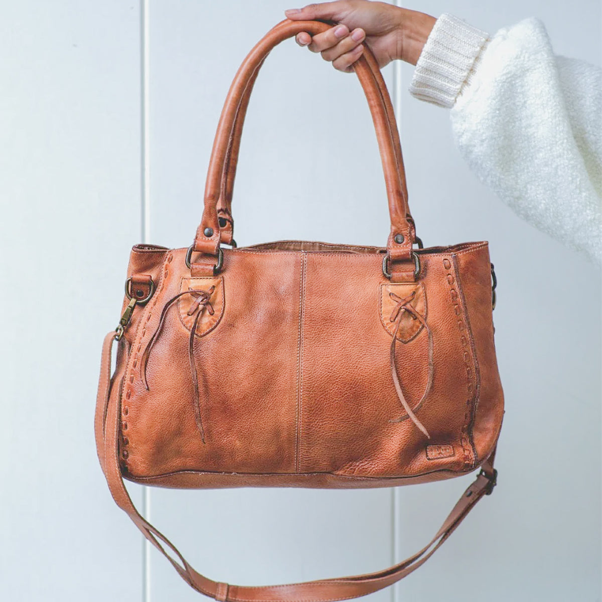 A hand holds the Rockababy by Bed|Stü—a compact leather bag crafted from vegetable-tanned leather, with dual handles, decorative stitching, and an attached crossbody strap—set against a plain white background.