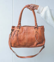 A hand holds the Rockababy by Bed|Stü—a compact leather bag crafted from vegetable-tanned leather, with dual handles, decorative stitching, and an attached crossbody strap—set against a plain white background.