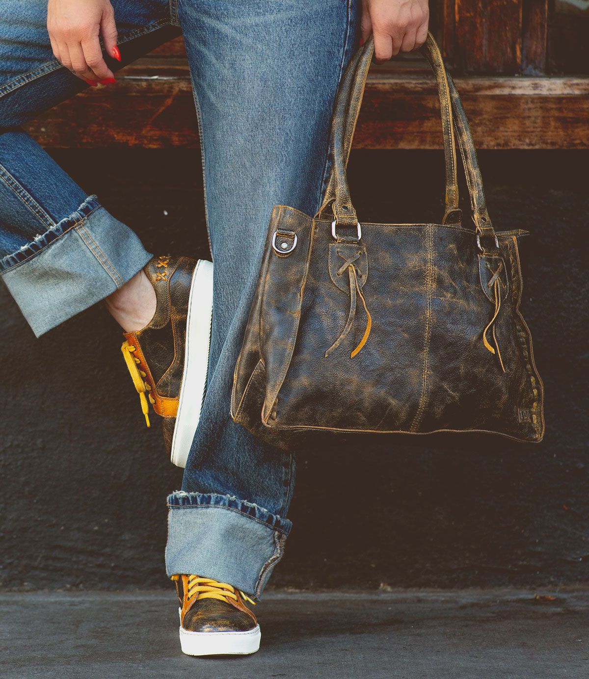 Wearing rolled-up jeans and sneakers, a person leans against a wooden surface with one foot crossed over the other, holding the Bed|Stü Rockababy—a compact bag crafted from vegetable-tanned leather.