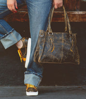 Wearing rolled-up jeans and sneakers, a person leans against a wooden surface with one foot crossed over the other, holding the Bed|Stü Rockababy—a compact bag crafted from vegetable-tanned leather.