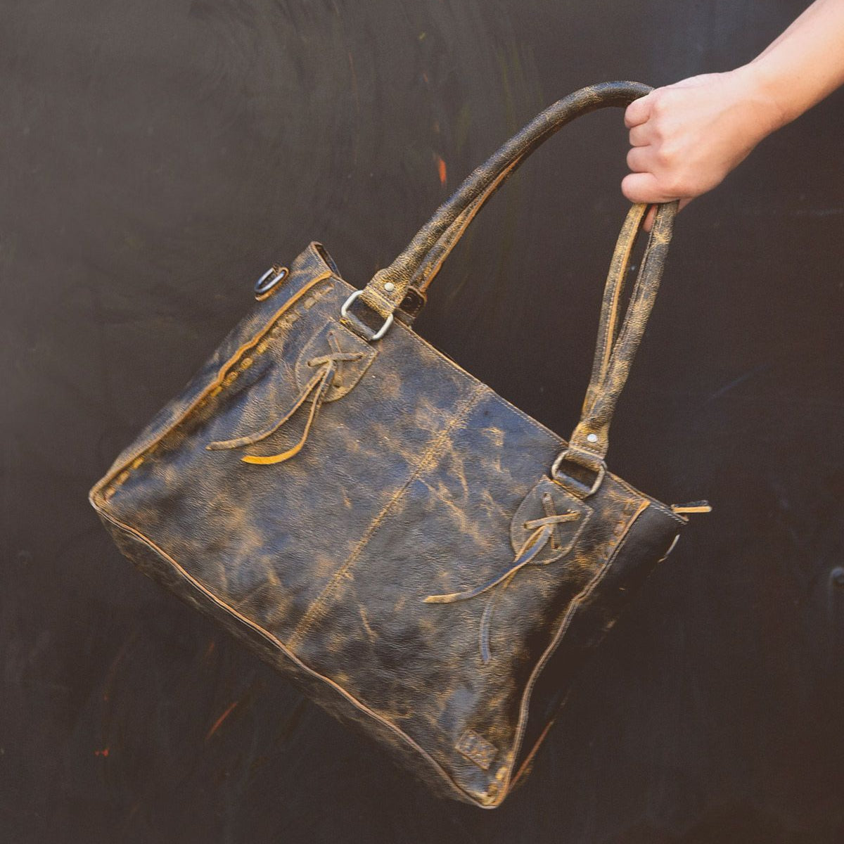 A hand holds the Rockababy by Bed|Stü, a compact leather bag crafted from distressed brown vegetable-tanned leather with double handles, metal hardware, and visible stitching, set against a dark background.