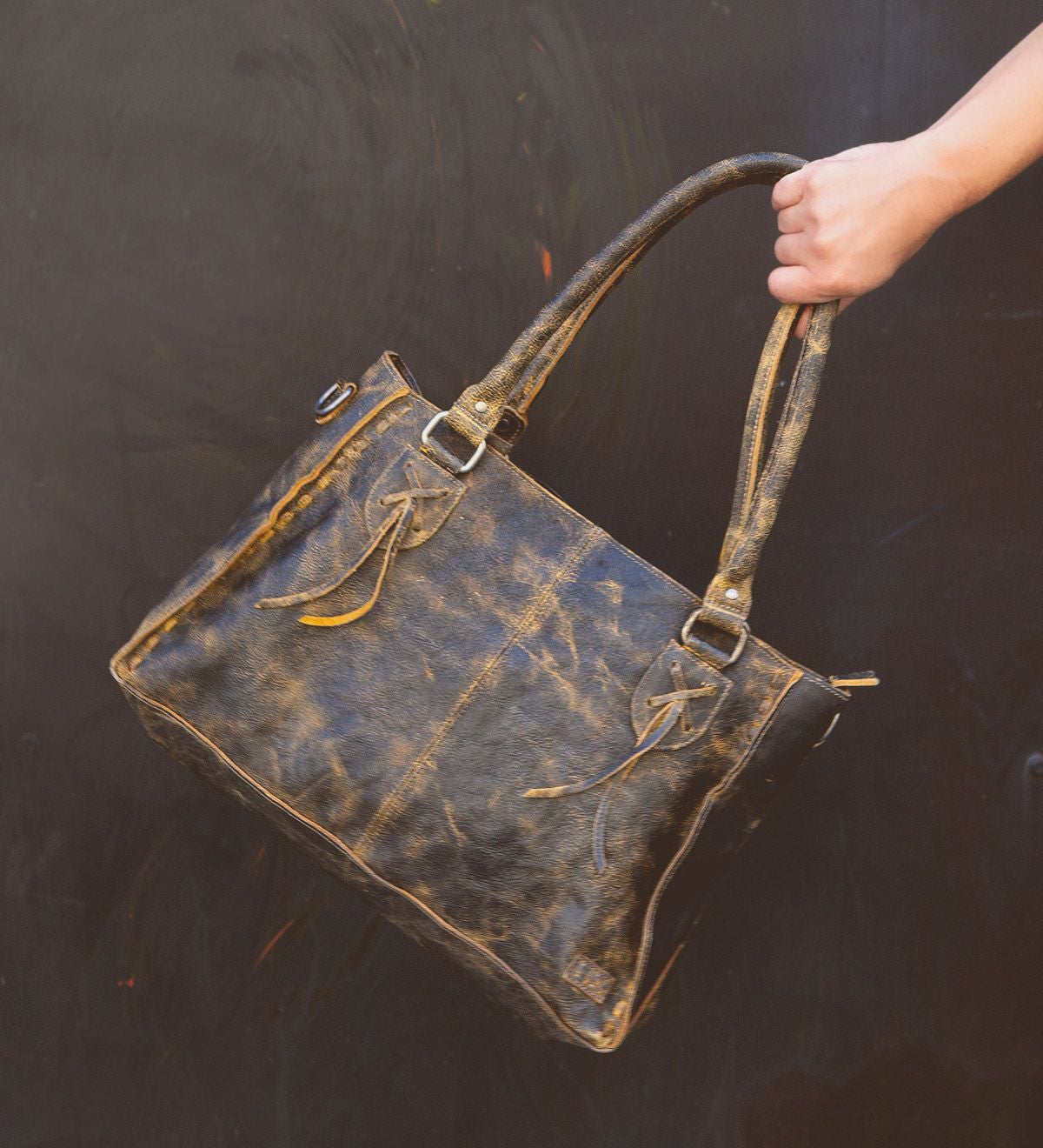 A hand holds the Rockababy by Bed|Stü, a compact leather bag crafted from distressed brown vegetable-tanned leather with double handles, metal hardware, and visible stitching, set against a dark background.