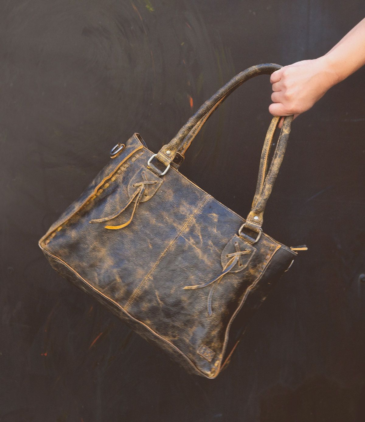 A hand holds the Rockababy by Bed|Stü, a compact leather bag crafted from distressed brown vegetable-tanned leather with double handles, metal hardware, and visible stitching, set against a dark background.