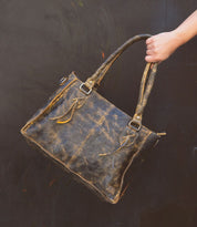 A hand holds the Rockababy by Bed|Stü, a compact leather bag crafted from distressed brown vegetable-tanned leather with double handles, metal hardware, and visible stitching, set against a dark background.