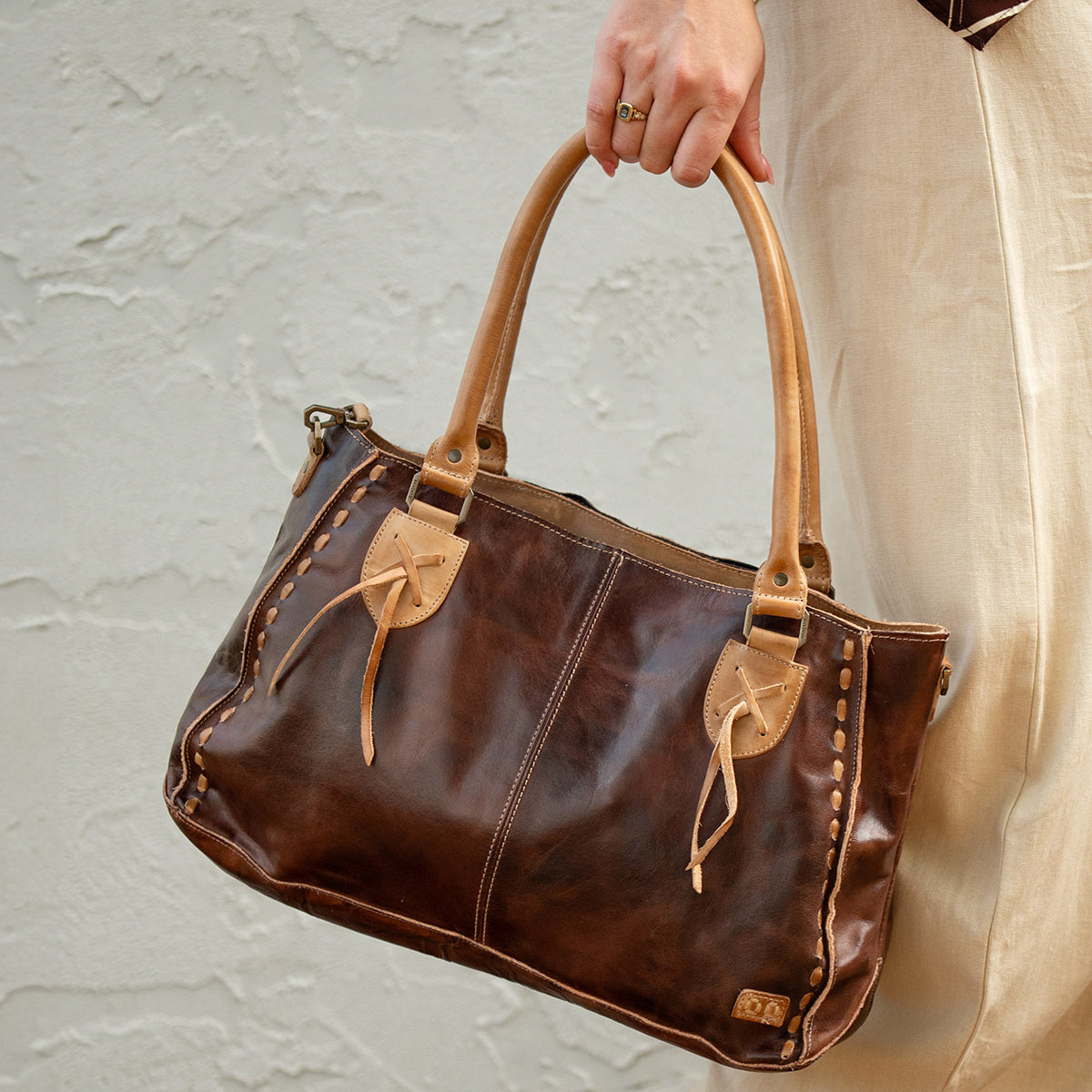 A person in beige pants holds the Bed|Stü Rockababy, a compact leather bag crafted from vegetable-tanned leather, featuring tan handles and decorative stitching, against a light textured wall.