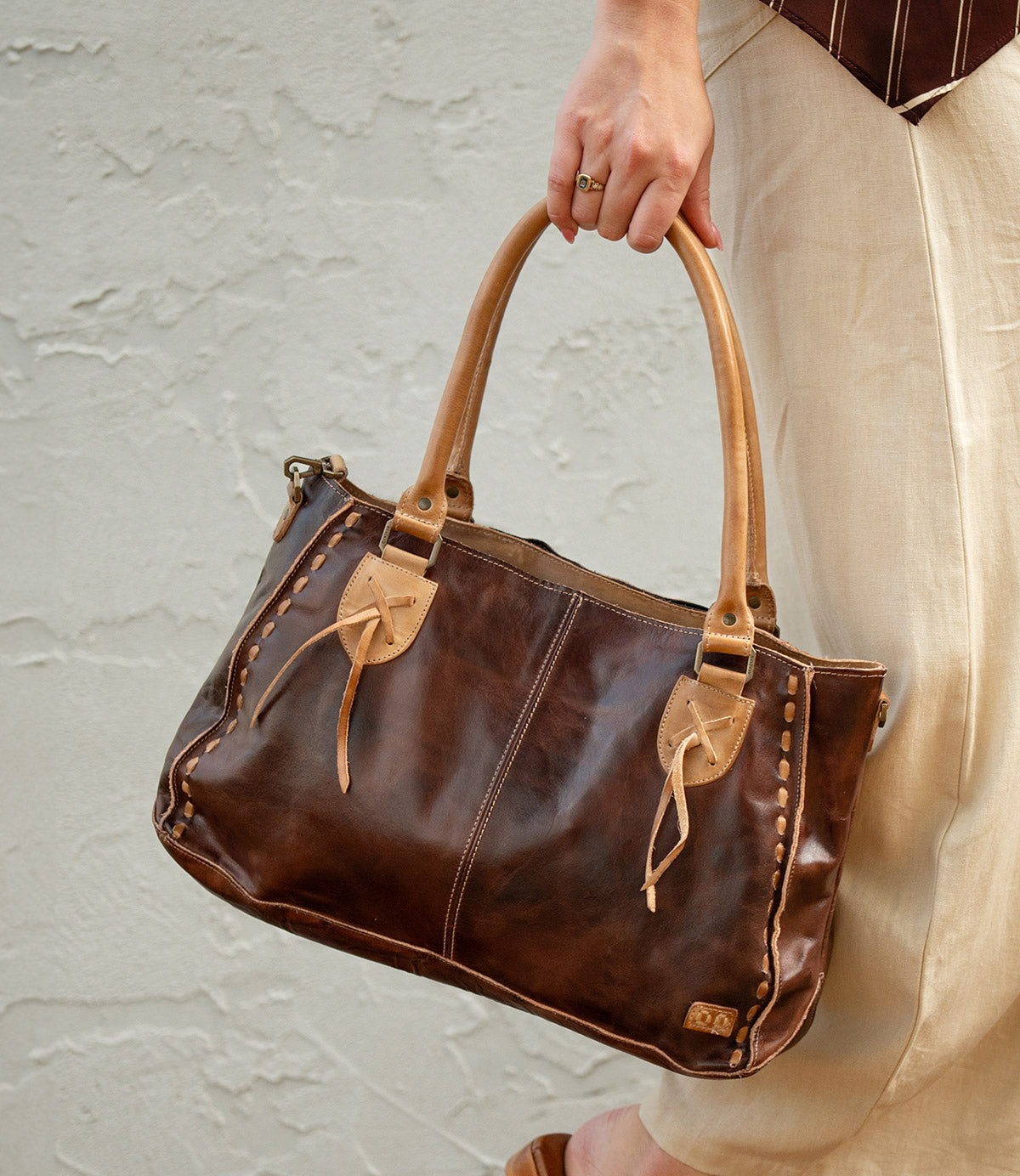 A person in beige pants holds the Bed|Stü Rockababy, a compact leather bag crafted from vegetable-tanned leather, featuring tan handles and decorative stitching, against a light textured wall.