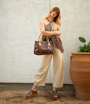 A woman stands in front of a textured wall, wearing a striped halter top, beige pants, platform sandals, and holding the Bed|Stü Rockababy compact leather bag with a crossbody strap.