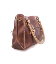 The Bed|Stü Rockababy is a brown, vegetable-tanned leather handbag featuring tan handles, visible stitching details, and a zipper pocket on the side, photographed on a white background. This compact leather bag also includes a convenient crossbody strap for versatile carrying options.