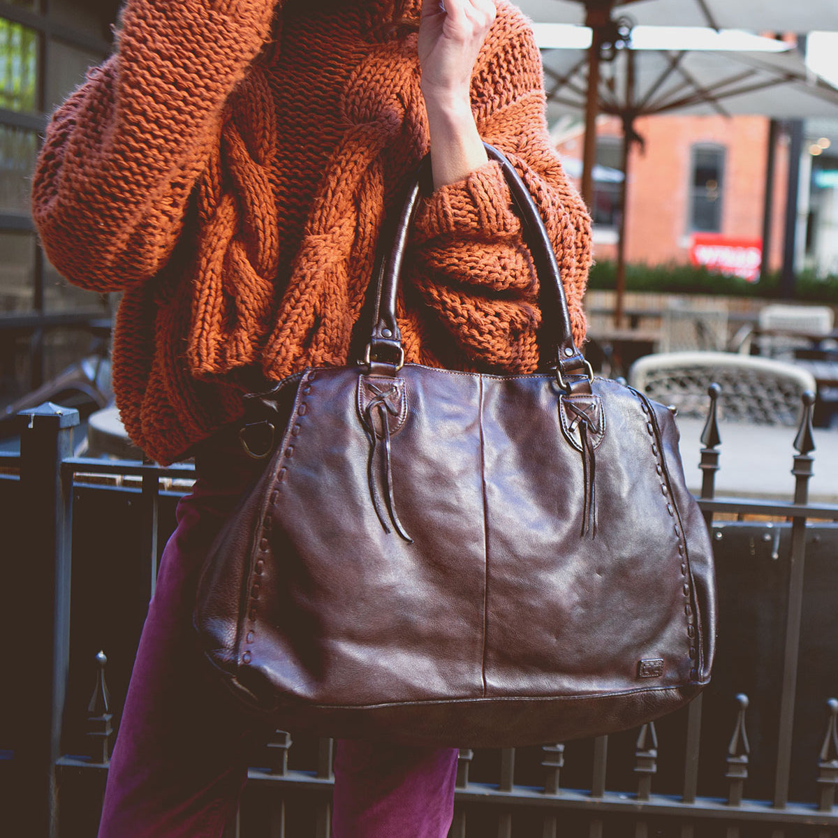 A person in an orange knit sweater holding the large brown full-grain leather Rockaway handbag by Bed|Stü, standing outdoors near patio tables and umbrellas.