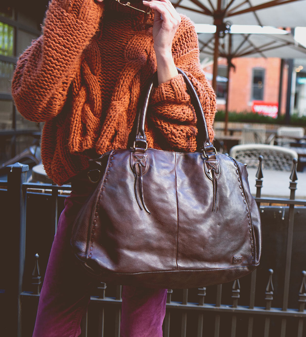 A person in an orange knit sweater holding the large brown full-grain leather Rockaway handbag by Bed|Stü, standing outdoors near patio tables and umbrellas.