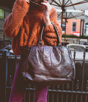 A person in an orange knit sweater holding the large brown full-grain leather Rockaway handbag by Bed|Stü, standing outdoors near patio tables and umbrellas.