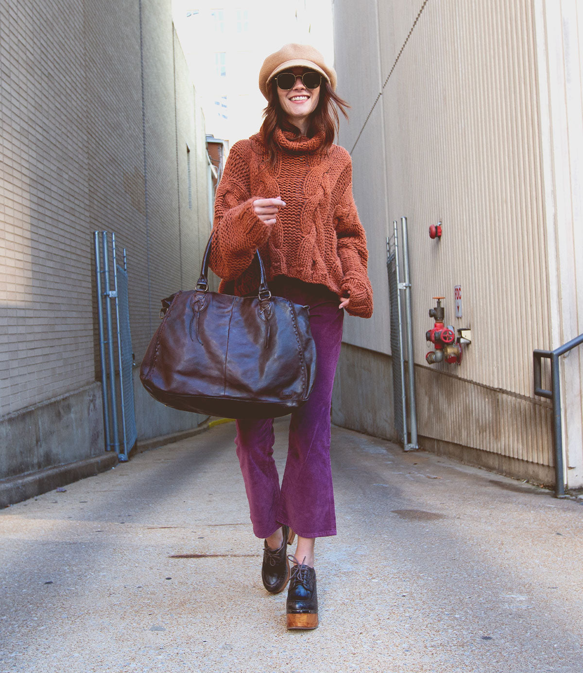 A woman wearing a hat, sunglasses, orange sweater, and purple pants walks in an alley, smiling as she carries the Bed|Stü Rockaway—a large black bag crafted from full-grain leather for everyday function.