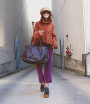A woman wearing a hat, sunglasses, orange sweater, and purple pants walks in an alley, smiling as she carries the Bed|Stü Rockaway—a large black bag crafted from full-grain leather for everyday function.