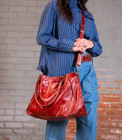 A person wearing a blue striped shirt and blue jeans stands against a brick wall, holding the Bed|Stü Rockaway—a large brown full-grain leather handbag with a shoulder strap.