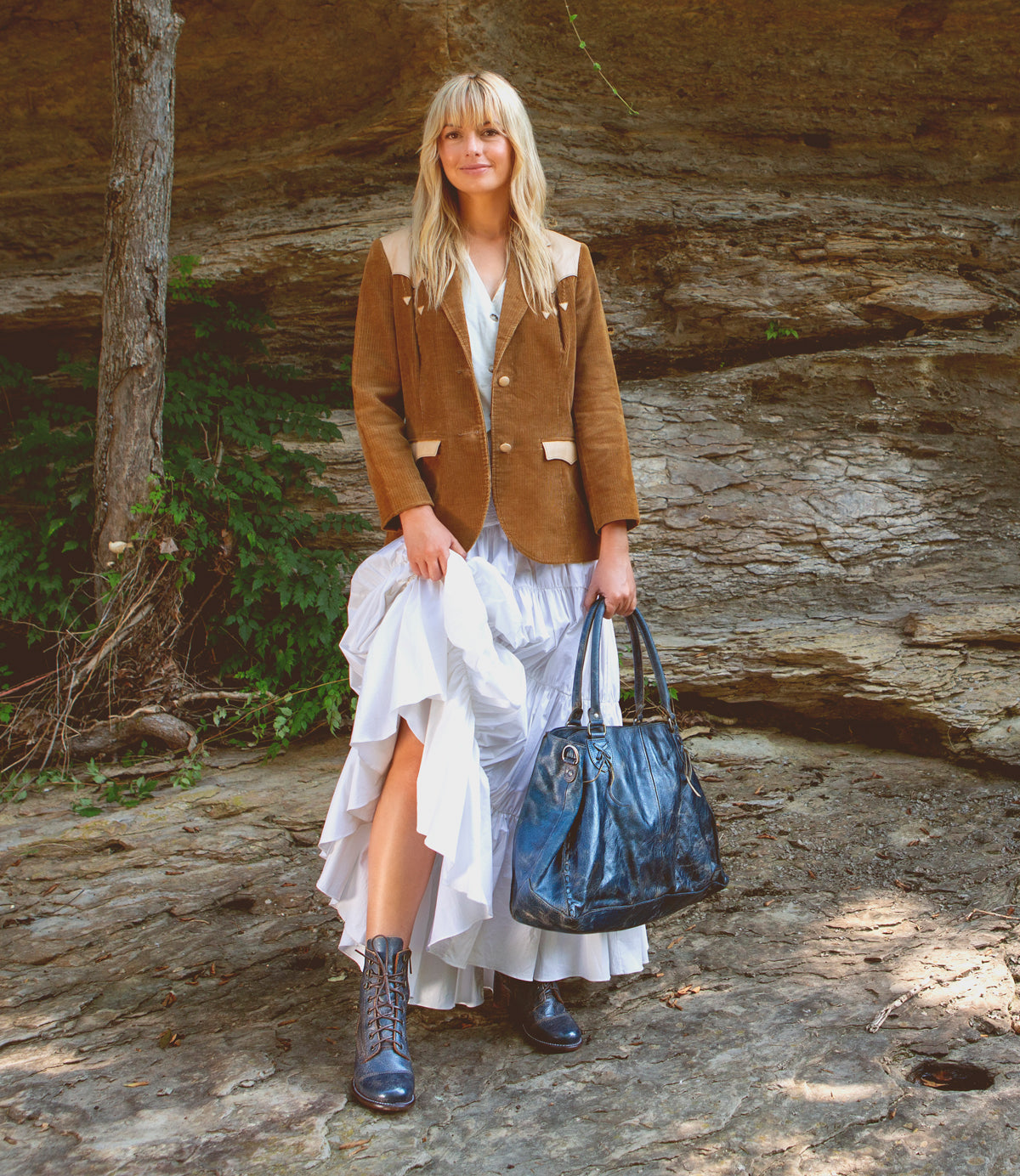 A woman stands outdoors on rocky ground wearing a brown Bed|Stü Rockaway vegetable-tanned leather jacket over a white ruffled dress, blue boots, and carrying a large blue bag.