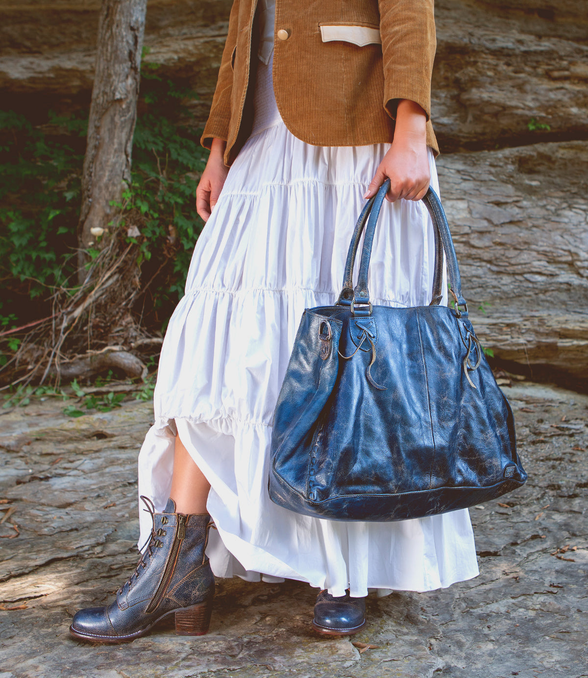 A person stands outdoors on rocks, wearing a white tiered skirt, brown boots, and a tan jacket, holding the Rockaway large black handbag by Bed|Stü, expertly crafted from vegetable-tanned leather.