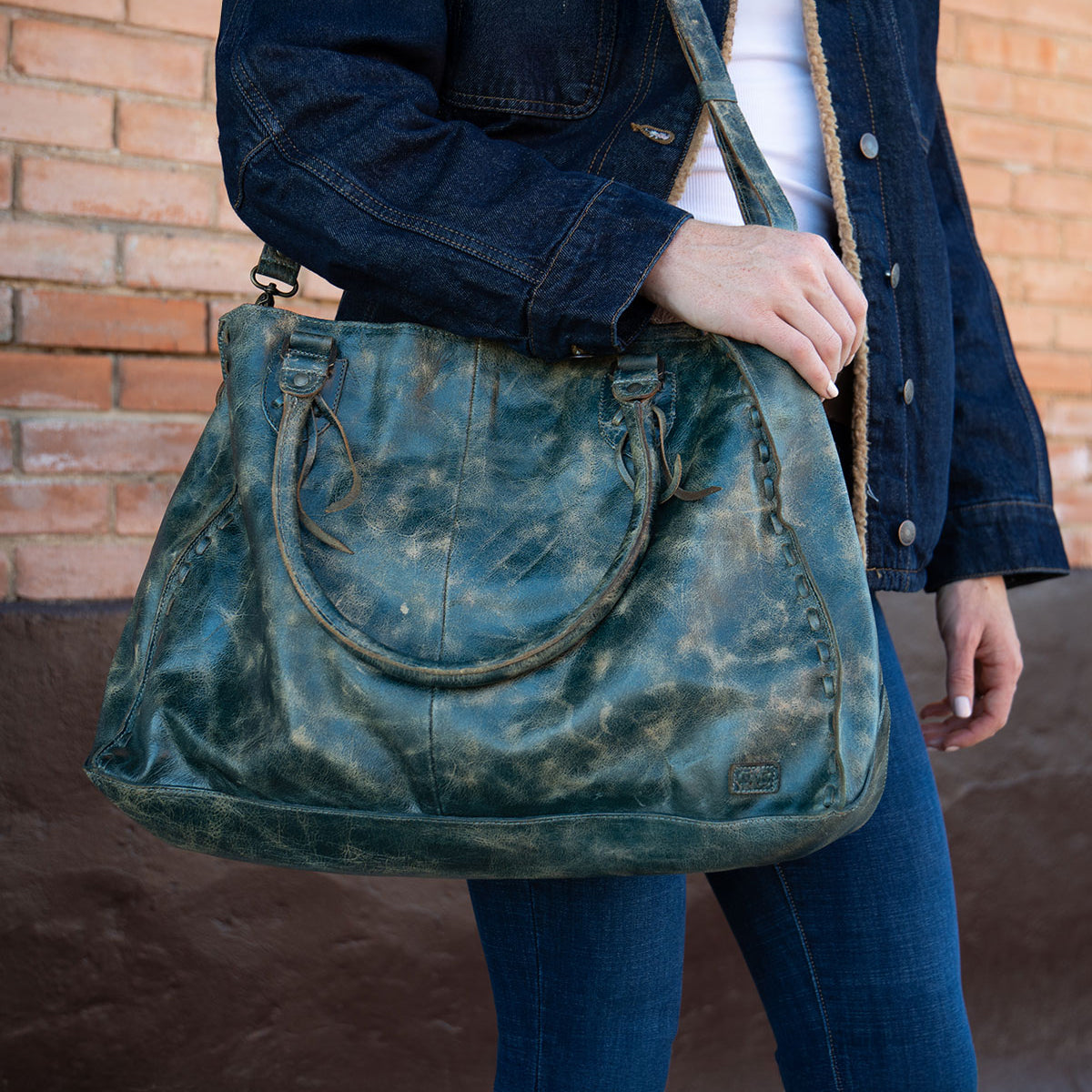 A person wearing a dark denim jacket and blue jeans holds the Bed|Stü Rockaway—a large, distressed green handbag crafted from full-grain leather, featuring two short handles and a shoulder strap—while standing in front of a brick wall. This piece beautifully blends everyday function with timeless style.