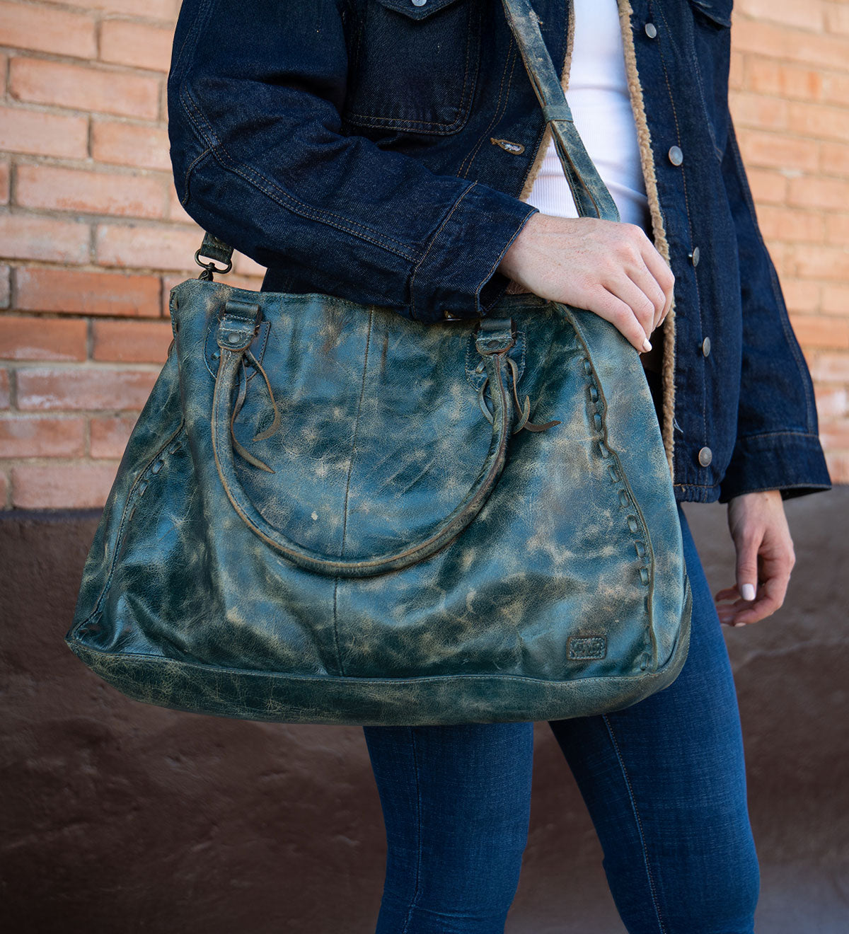 A person wearing a dark denim jacket and blue jeans holds the Bed|Stü Rockaway—a large, distressed green handbag crafted from full-grain leather, featuring two short handles and a shoulder strap—while standing in front of a brick wall. This piece beautifully blends everyday function with timeless style.