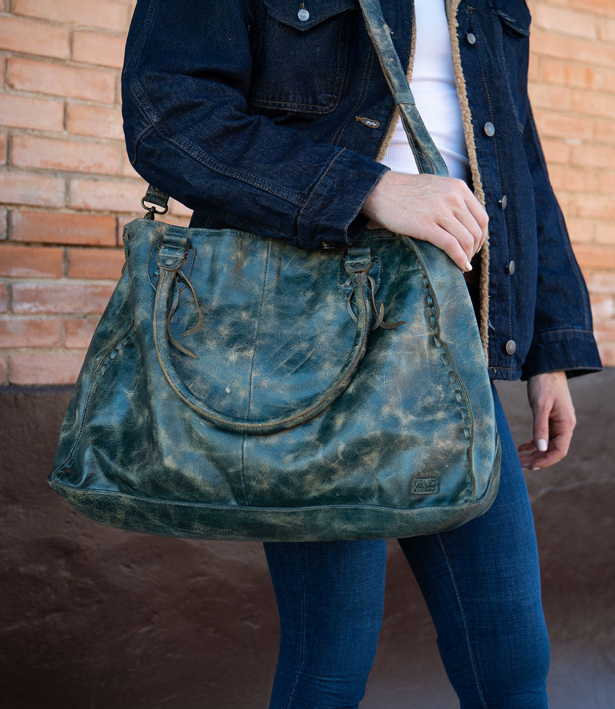 A person wearing a dark denim jacket and blue jeans holds the Bed|Stü Rockaway—a large, distressed green handbag crafted from full-grain leather, featuring two short handles and a shoulder strap—while standing in front of a brick wall. This piece beautifully blends everyday function with timeless style.