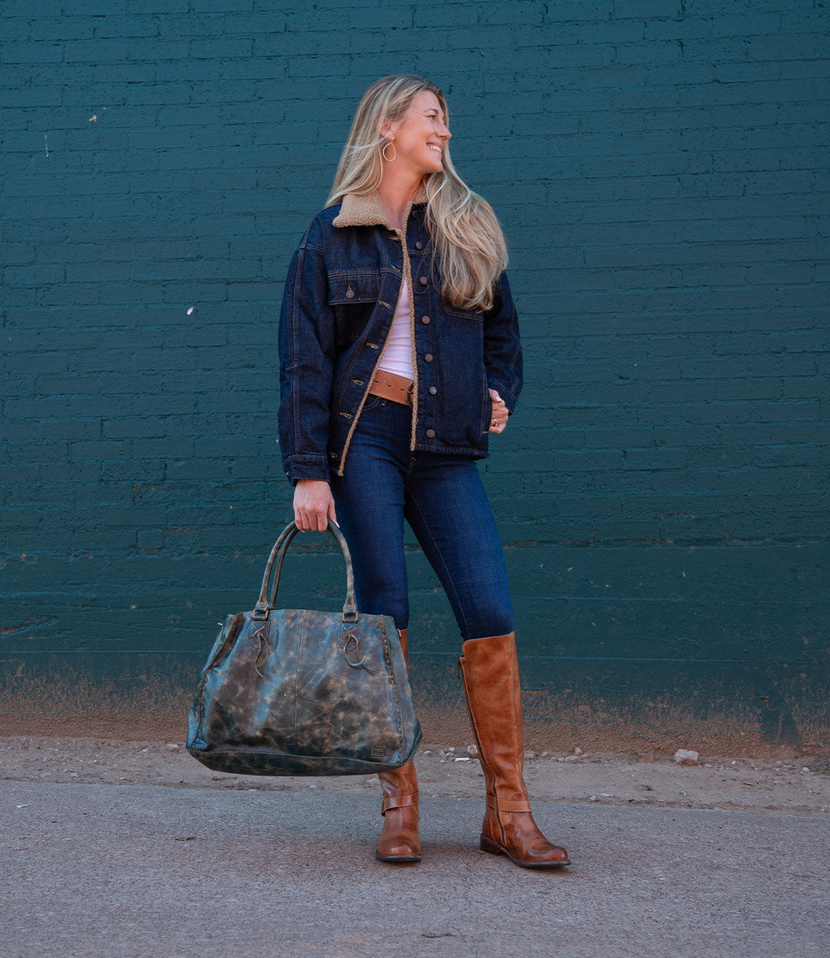 A woman stands against a green brick wall, wearing a denim jacket, blue jeans, and brown boots. She smiles as she looks to her left, holding the Bed|Stü Rockaway—a large green bag crafted from full-grain leather that’s perfect for everyday use.