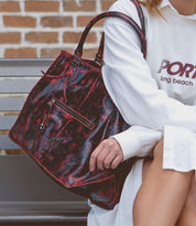 A person in a white long-sleeve shirt sits on a bench holding the Rockaway, a large black and red patterned full-grain leather handbag by Bed|Stü, perfectly balancing style with everyday function.
