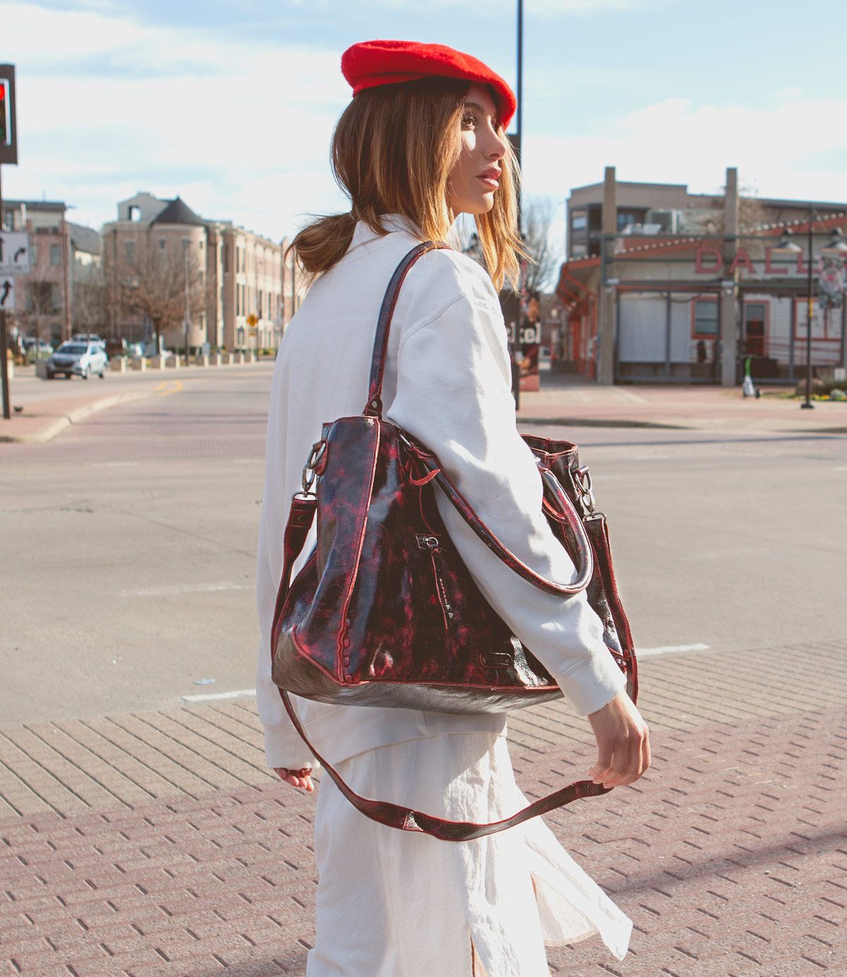 A woman in a red beret and white coat stands on a city street, carrying the Bed|Stü Rockaway—a large maroon full-grain leather bag—over her shoulder, looking away from the camera.