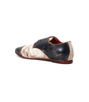 The Bed|Stü Rumba II is a two-tone lace-up leather shoe featuring scuffed black and beige leather, a red lining, and a flat sole. Shown angled from the back on a white background, this shoe effortlessly blends vintage charm with stylish comfort.