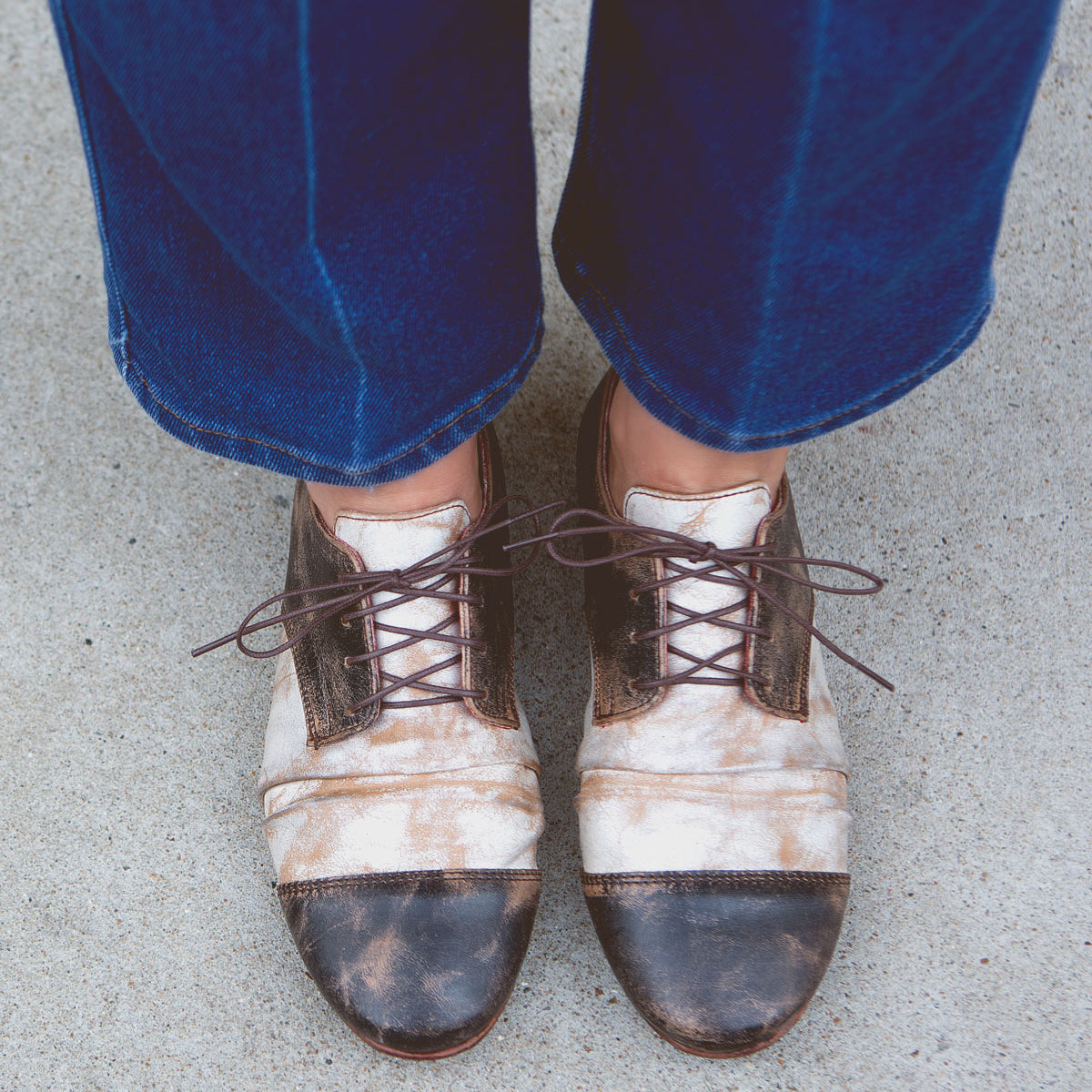 A person wearing Bed|Stü Rumba II brown and white distressed lace-up leather shoes with blue jeans stands on a concrete surface, viewed from above.