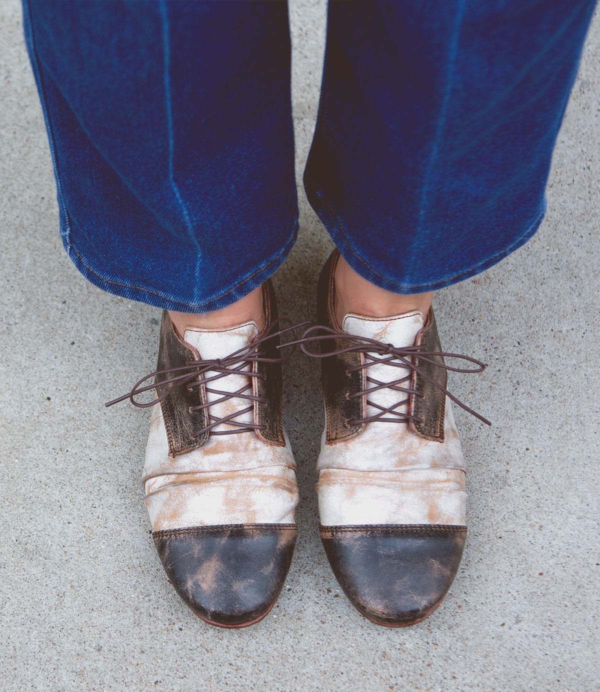 A person wearing Bed|Stü Rumba II brown and white distressed lace-up leather shoes with blue jeans stands on a concrete surface, viewed from above.