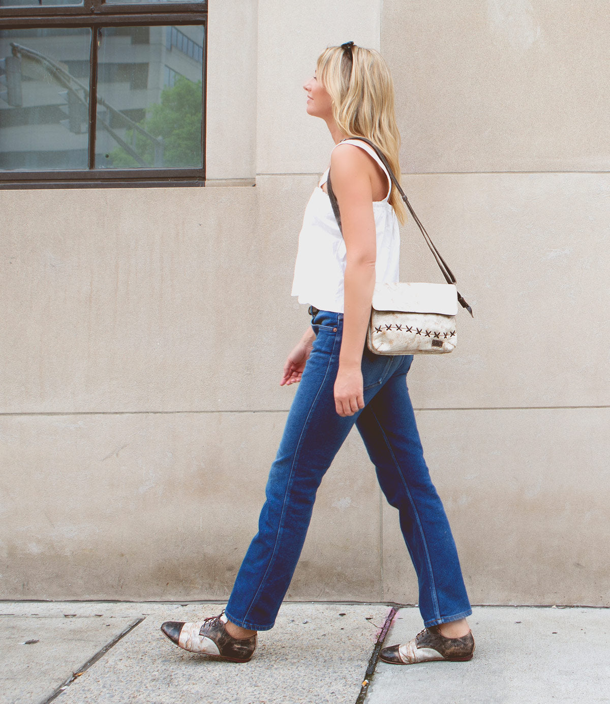 A woman with blonde hair walks past a building wearing a white top, blue jeans, the Bed|Stü Rumba II stylish comfort lace-up leather shoes, and a crossbody bag.