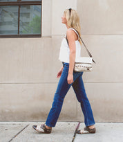 A woman with blonde hair walks past a building wearing a white top, blue jeans, the Bed|Stü Rumba II stylish comfort lace-up leather shoes, and a crossbody bag.