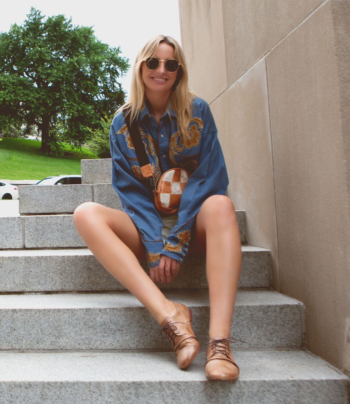 A blonde woman wearing a blue patterned shirt and Bed|Stü Rumba II stylish comfort lace-up leather shoes sits on stone stairs outdoors.