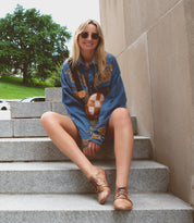 A blonde woman wearing a blue patterned shirt and Bed|Stü Rumba II stylish comfort lace-up leather shoes sits on stone stairs outdoors.
