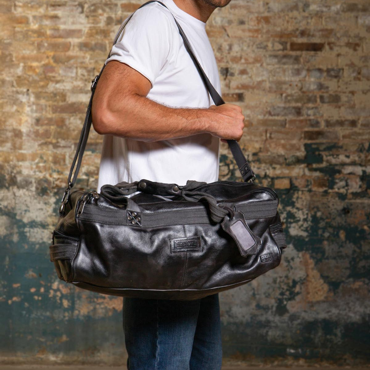 A man in a white t-shirt and jeans carries the Bed|Stü Ruslan large black leather duffle travel bag with durable canvas straps, standing against a worn brick wall.