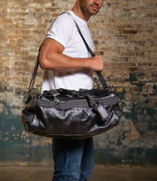 A man in a white t-shirt and jeans carries the Bed|Stü Ruslan large black leather duffle travel bag with durable canvas straps, standing against a worn brick wall.
