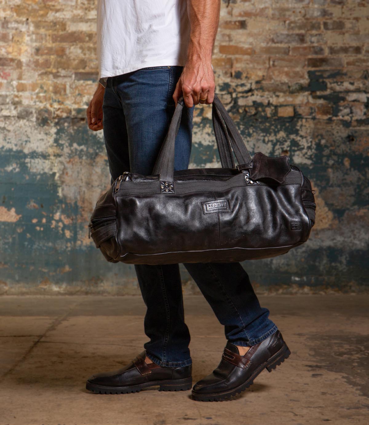 A man in jeans and loafers holds the Ruslan black leather duffel bag by Bed|Stü, featuring durable canvas straps, while standing indoors against a worn brick and painted wall.