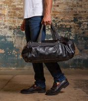 A man in jeans and loafers holds the Ruslan black leather duffel bag by Bed|Stü, featuring durable canvas straps, while standing indoors against a worn brick and painted wall.