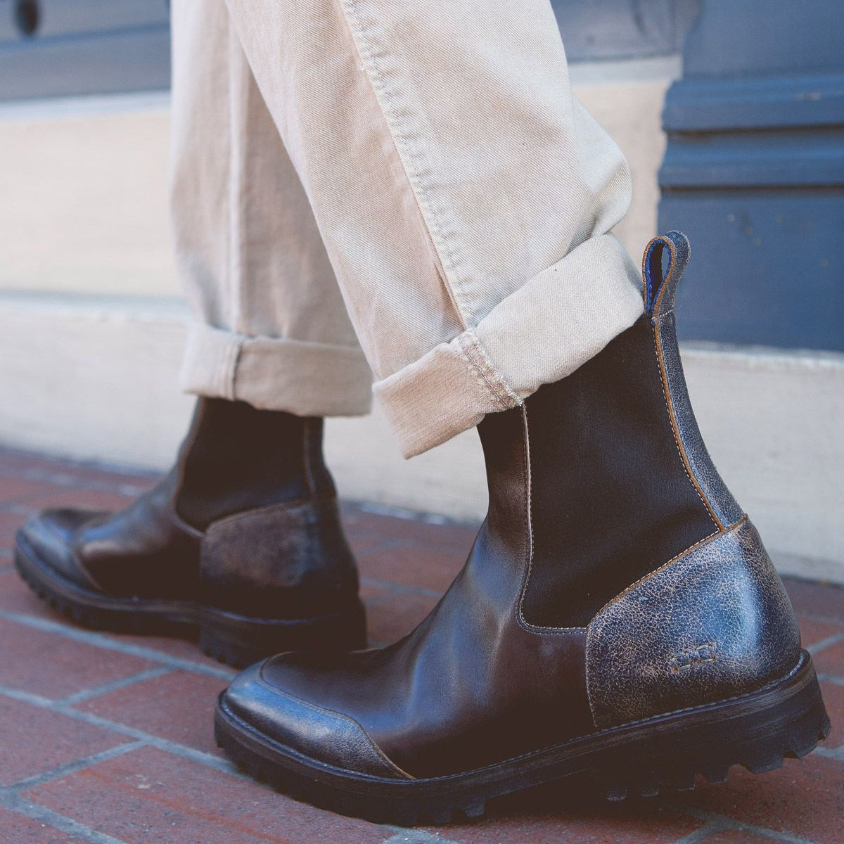 A person wearing cuffed beige pants and the Bed|Stü Salatiel dark brown leather Chelsea boots with a cushioned leather footbed stands on a brick sidewalk next to a blue wall.