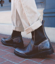 A person wearing cuffed beige pants and the Bed|Stü Salatiel dark brown leather Chelsea boots with a cushioned leather footbed stands on a brick sidewalk next to a blue wall.