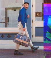 A man wearing a blue shirt, beige pants, and stylish Bed|Stü Salatiel Chelsea boots with a textured outsole walks on a sidewalk, holding a brown leather duffel bag.