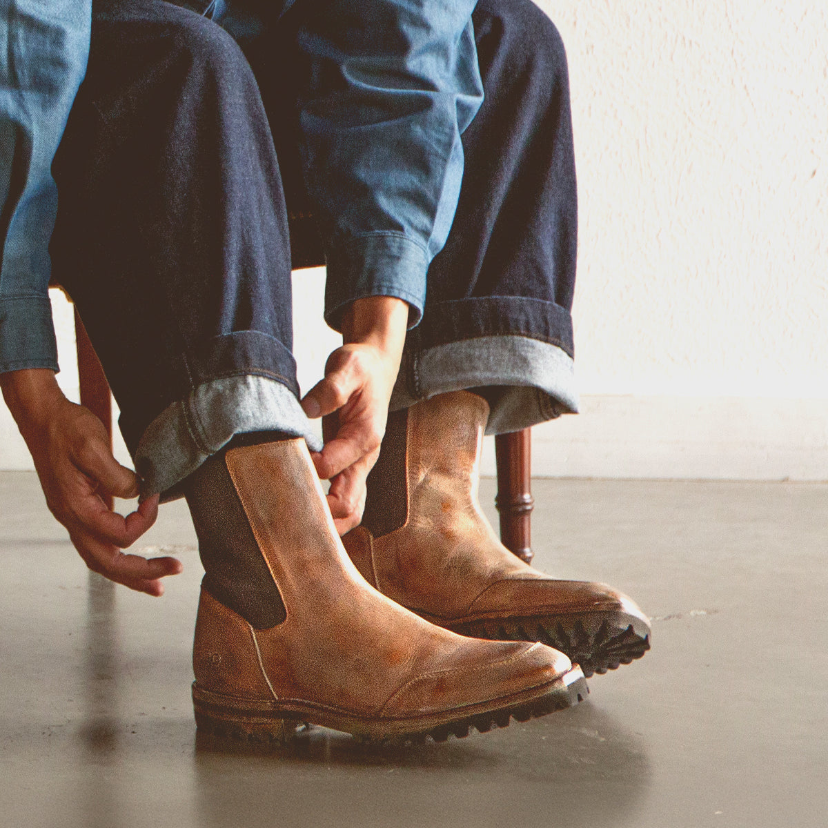A person sits indoors on a simple wooden chair, adjusting the cuffs of their blue jeans while wearing the Bed|Stü Salatiel light brown leather Chelsea boots, which feature a textured outsole and cushioned leather footbed, paired with a denim jacket.