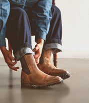 A person sits indoors on a simple wooden chair, adjusting the cuffs of their blue jeans while wearing the Bed|Stü Salatiel light brown leather Chelsea boots, which feature a textured outsole and cushioned leather footbed, paired with a denim jacket.