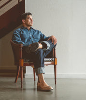 A man sits in a wooden chair with his legs crossed, wearing a denim shirt, rolled-up jeans, and Bed|Stü’s Salatiel brown slip-on shoes featuring a textured outsole, gazing to the side in a minimalist indoor setting.