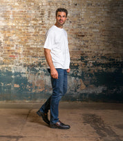 A man in a white T-shirt, blue jeans, and Bed|Stü's Samosa loafers stands sideways in front of a worn brick wall, looking toward the camera.