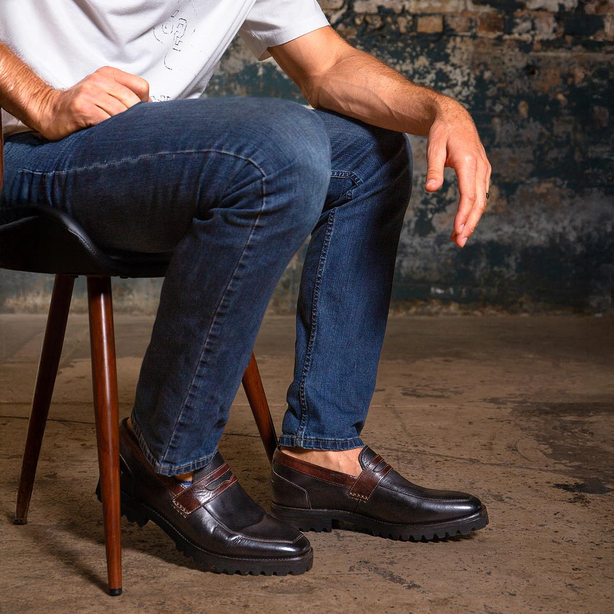 A seated person wearing a white shirt, blue jeans, and Bed|Stü Samosa classic loafers with thick soles, set against a rough, industrial background.