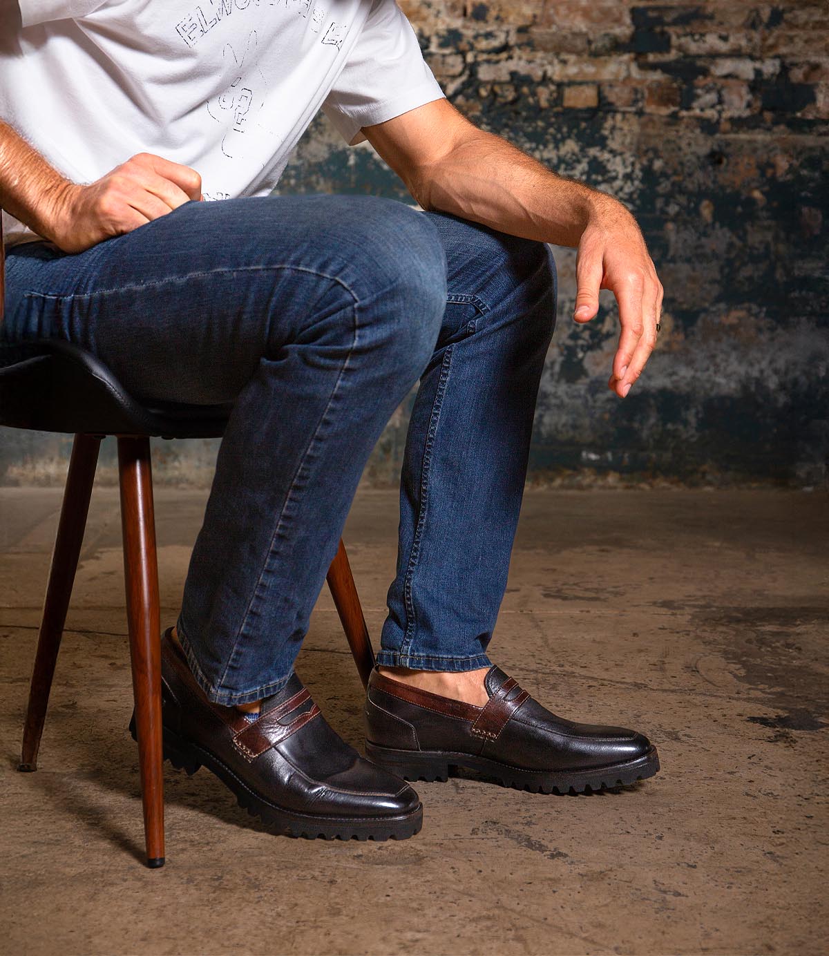 A seated person wearing a white shirt, blue jeans, and Bed|Stü Samosa classic loafers with thick soles, set against a rough, industrial background.