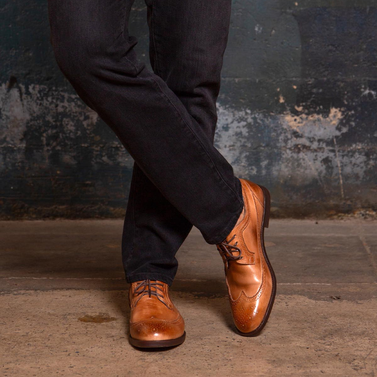 A person wearing dark pants and the Bed|Stü Sandro II tan handcrafted leather dress shoes with elegant wingtip broguing stands with legs crossed on a concrete floor against a textured wall.