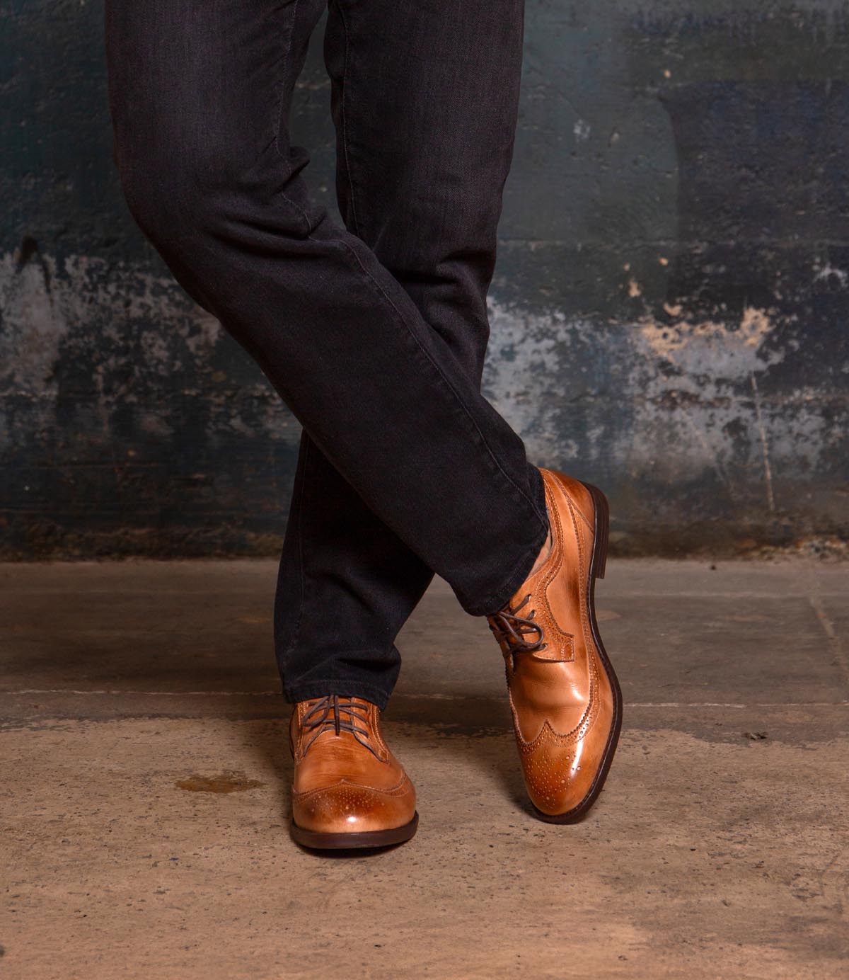 A person wearing dark pants and the Bed|Stü Sandro II tan handcrafted leather dress shoes with elegant wingtip broguing stands with legs crossed on a concrete floor against a textured wall.