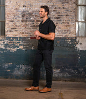 A man stands smiling in front of an old brick wall, showcasing timeless style in a black short-sleeve shirt, black pants, and Bed|Stü’s Sandro II handcrafted leather dress shoes featuring elegant wingtip broguing.